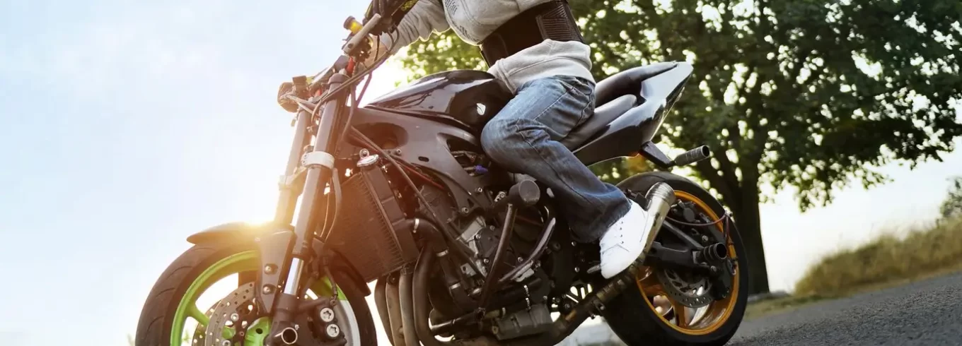 Motociclista con casco y ropa de protección montando una moto deportiva negra en una carretera al aire libre, rodeado de árboles y luz natural.